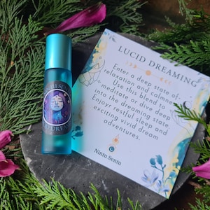 Può includere: Un flacone roll-on in vetro turchese con tappo blu, etichettato "Nintu Sentu Lucid Dream". Una carta con testo sui sogni lucidi è accanto al flacone. L'immagine è circondata da verde e petali di fiori rosa.