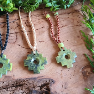 May include: Three green stone pendant necklaces with a cross-like design, each suspended from a different cord: black, beige, and brown. The pendants have a circular hole in the centre. The necklaces are displayed on a wooden surface with green foliage.