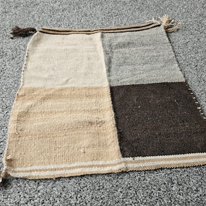 May include: A square woven textile piece featuring a colour-blocked design in cream, light grey, tan, and dark brown. The fabric has a visible coarse texture with horizontal striped borders at the top and bottom edges, finished with small knotted fringe tassels.
