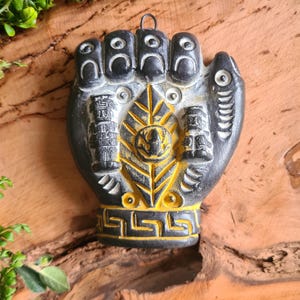 Peut inclure: Décoration murale en forme de main noire et dorée avec des motifs complexes. La main présente des sculptures détaillées d'éléments architecturaux et de motifs géométriques. Sur fond de bois naturel, avec du feuillage vert.