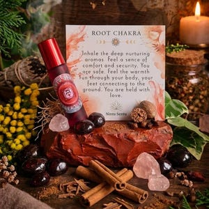 May include: A red essential oil roller bottle with a label, resting on a red rock with cinnamon sticks, crystals, and other natural elements. A card reads "ROOT CHAKRA" with text about grounding and comfort.