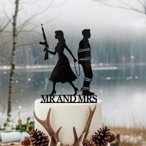 May include: A two-tiered wedding cake with a burlap-wrapped base, decorated with pine cones and antlers. A black silhouette cake topper depicts a bride with a rifle and a groom tied up. The words "MR AND MRS" are below the topper.