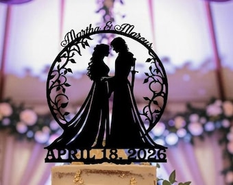 May include: A three-tiered wedding cake with white frosting and floral decorations. A black silhouette cake topper features two figures and the names "Martha & Marcus" with the date "April 18, 2026". The cake sits on a wooden cake stand.