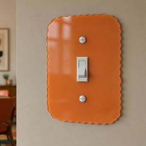 May include: An orange light switch cover with a scalloped edge and a white toggle switch. The cover is mounted on a beige wall. The image also shows a dining room with a round wooden table, orange chairs, and a patterned rug.