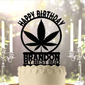 Può includere: Un cake topper nero con le parole "HAPPY BIRTHDAY" e un disegno a forma di foglia di cannabis. Sotto la foglia c'è il testo "BRANDON MY BEST BUD". La torta bianca ha un motivo a foglie e poggia su una base di legno.