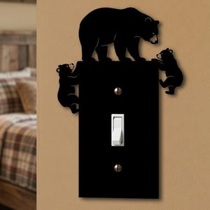 Pode incluir: Uma placa de cobertura de interruptor de luz retangular preta com a silhueta de um urso grande em cima e dois filhotes de urso subindo pelas laterais. A placa inclui um interruptor de alavanca branco e dois parafusos de montagem prateados.