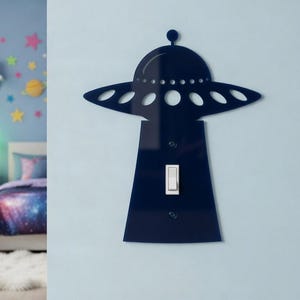 Può includere: Una copertura per interruttore della luce in acrilico blu navy a forma di UFO. La copertura ha una parte superiore circolare con ritagli ovali e una base rettangolare. L'interruttore della luce è bianco. Lo sfondo è una parete azzurra.