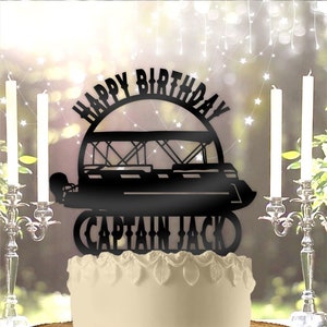 May include: Black silhouette cake topper with the text "Happy Birthday" in a circle above a silhouette of a pontoon boat and the text "Captain Jack" below. The topper is on a white cake with a wooden cake stand.