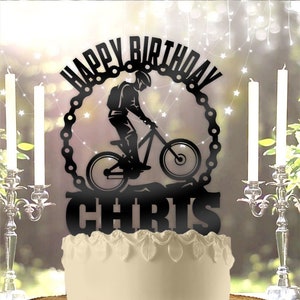 May include: Black cake topper with a silhouette of a person riding a mountain bike. The topper says "Happy Birthday Chris" in a circle with a chain design.