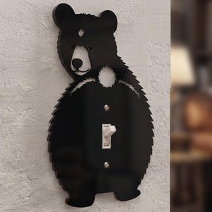 May include: A black bear-shaped light switch cover plate featuring a cutout design of a bear's head and body. The plate is made of a glossy black material and includes a central rectangular opening for a standard toggle light switch.