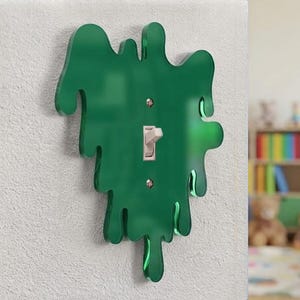 Könnte beinhalten: Eine dunkelgrüne Lichtschalterabdeckung mit einer glänzenden, unregelmäßigen, tropfenartigen Silhouette. Die Abdeckung besteht aus durchscheinendem Acrylmaterial und ist mit zwei silberfarbenen Metallschrauben um einen weißen Kippschalter befestigt.