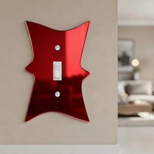 May include: A decorative red light switch cover with a unique, stylized shape. The cover has a glossy finish and features a standard light switch and two silver screws. The switch is mounted on a neutral-colored wall.
