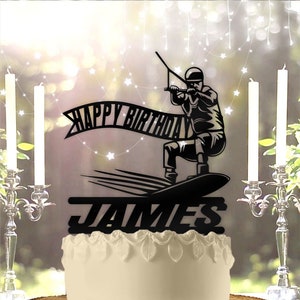 May include: Black silhouette cake topper of a person wakeboarding with a banner that says "Happy Birthday" and the name "JAMES" underneath.