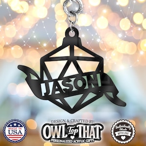 May include: Black acrylic ornament shaped like a 20-sided die with the name "JASON" inscribed on it. The ornament is hanging from a silver chain. The ornament is against a blurry background of twinkling lights.