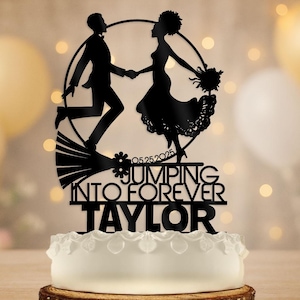 May include: Black silhouette cake topper featuring a running couple holding hands, with the words "Jumping Into Forever Taylor" and the date 05.25.2025. The topper is placed on a white cake with decorative frosting.