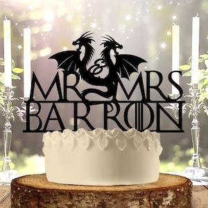 Può includere: Silhouette nera di due draghi con le ali spiegate, che formano un cuore con le loro code. Il testo "MR & MRS BARRON" è sotto i draghi. Il topper per torta è su una torta bianca con una base di legno.