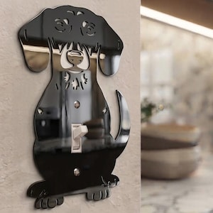 Puede incluir: Una placa de interruptor de luz negra y reflectante con forma de perro. El diseño presenta una silueta recortada de un perro con orejas caídas, cola y patas. Un interruptor de palanca blanco se sitúa en el centro, fijado con dos tornillos.