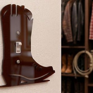 May include: A dark brown, glossy, cowboy boot-shaped light switch wall plate. The plate features a cutout for a single toggle switch and two circular holes for mounting screws, with a smooth, reflective surface and defined boot silhouette.