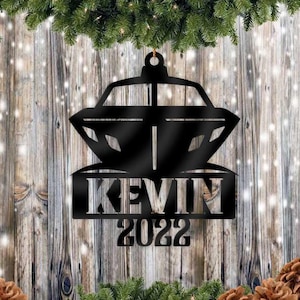 Puede incluir: Adorno navideño de metal negro con el nombre "KEVIN" y el año "2022" recortados. El adorno tiene una forma decorativa y está colgado sobre un fondo de madera con agujas de pino y piñas.