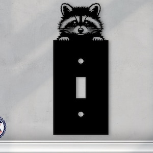 May include: Black metal light switch cover with a raccoon peeking over the top. The switch cover has a single toggle switch and two screw holes.