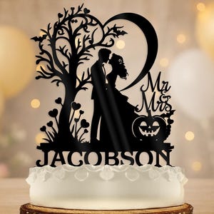 Puede incluir: Adorno negro para pastel con la silueta de una pareja besándose bajo un árbol y un corazón. Incluye "Mr & Mrs" y una calabaza de Halloween. El apellido "Jacobson" se muestra en la parte inferior. El pastel es blanco con borde decorativo.