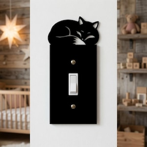 May include: A black rectangular light switch cover plate featuring a decorative cutout of a sleeping fox at the top. The plate is secured to a white wall with two visible metal screws and contains a single white vertical toggle light switch.