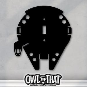 May include: Black acrylic light switch cover in the shape of the Millennium Falcon spaceship. The cover has a rectangular opening for the switch and several circular cutouts. The logo "OWL Top THAT" is at the bottom.