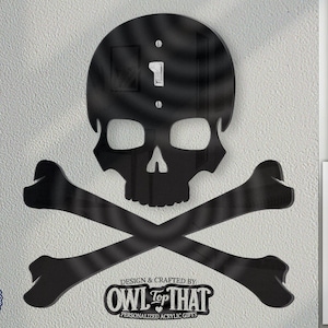 Op de afbeelding: Zwart schedel- en kruisbeendesign met een lichtschakelaar of stopcontact in het midden. Het ontwerp is gemaakt van acryl en is gepersonaliseerd met de tekst "OWL TOP THAT".