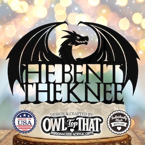 May include: Black acrylic cake topper featuring a dragon with wings spread out and the text "HE BENT THE KNEE". The topper is made by OWL TOP THAT, a company that makes personalized acrylic gifts.