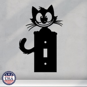 Op de afbeelding: Zwart-witte silhouet van een kat met een ondeugende grijns, geplaatst boven een lichtschakelaar. De staart van de kat wikkelt zich om de schakelaarplaat.