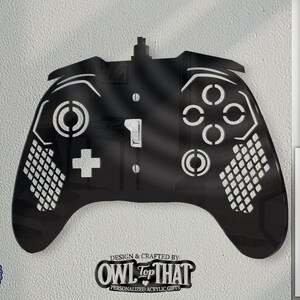 May include: Black acrylic wall art shaped like a video game controller. The controller features white detailing for buttons and directional pads. The design includes the text "DESIGN & CRAFTED BY OWL TOP THAT PERSONALIZED ACRYLIC GIFTS" and "MADE IN USA".