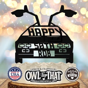 May include: Black silhouette of a DeLorean time machine with the words "Happy 52nd Rob" on the front. The silhouette is on top of a white cake with a red, white, and blue "Made in USA" sticker, a black and white "OWL Top THAT" logo, and a white "Individually Made with Love" sticker.