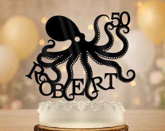 Puede incluir: Adorno negro para pastel con forma de pulpo con el nombre "ROBERT" y el número "50". El adorno está colocado sobre un pastel blanco con un borde decorativo, sobre una base de madera. El fondo está borroso con luces bokeh.