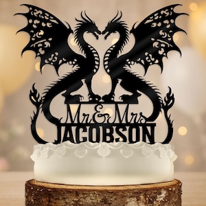 以下が含まれることがあります： 黒いドラゴンのケーキトッパー。「Mr & Mrs JACOBSON」の文字入り。 ドラゴンは互いに向き合い、ハート型を形成しています。 ケーキトッパーは、装飾的な縁のある白いケーキの上にあり、木の台座に置かれています。