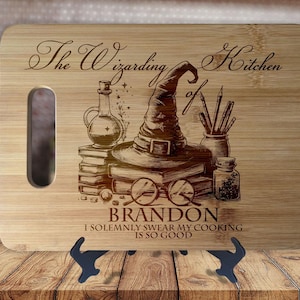 Może przedstawiać: Drewniana deska do krojenia z wygrawerowanym tekstem "The Wizarding Kitchen of Brandon I Solemnly Swear My Cooking Is So Good". Deska przedstawia kapelusz czarodzieja, butelkę z eliksirem i pióro.