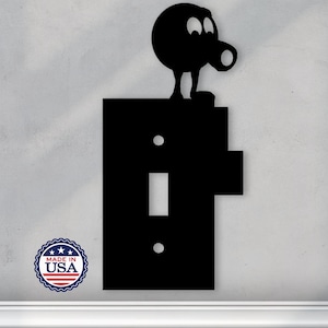May include: Black metal light switch cover with a black and white cartoon character on top. The character has a round head with two eyes and a mouth. The switch cover has a single toggle switch and two screw holes. The text "Made in USA" is printed on a small circle sticker.