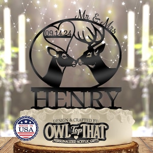 May include: Black metal wedding cake topper featuring two deer silhouettes with a heart and the text "Mr. & Mrs. 09.14.24 HENRY". The topper is on a white cake with a brown base and a sticker that says "Made in USA" and "Design & Crafted by: OWL Top THAT Personalised Acrylic Gifts".