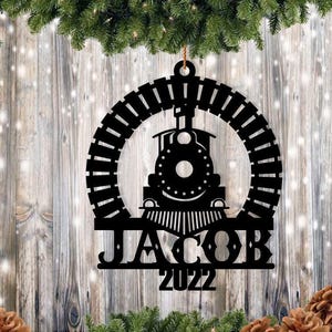 May include: Black metal train ornament with the name "JACOB" and the year "2022" cut out. The train is centered within a circular track design. The ornament is hanging from a brown string, with a wooden background and Christmas decorations.