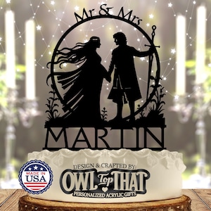 May include: Black acrylic cake topper featuring a silhouette of a couple holding hands within a circular frame, with the words "Mr & Mrs" above and "MARTIN" below. The cake topper is on a white cake with the text "DESIGN & CRAFTED BY: OWL TOP THAT".