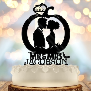 May include: Black acrylic wedding cake topper with a kissing couple silhouette inside a circular frame. The date "10.31.25" is above the couple, with "MR & MRS JACOBSON" below. A small bat is on the right side of the name.