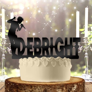 May include: A black cake topper with the name "DEBRIGHT" and a silhouette of a singer with a microphone. The cake sits on a wooden base, with a white cake and decorative candles in the background.