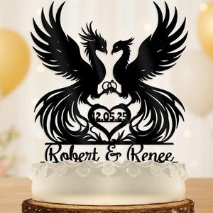May include: Black phoenix cake topper with the names "Robert & Renee" and the date "12.05.25" cut out. The topper features two phoenixes facing each other with their wings spread. The cake is white with a decorative edge, set on a wooden base.