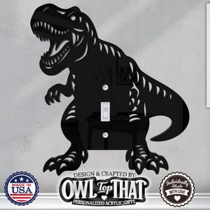 May include: Black Tyrannosaurus Rex light switch cover. The dinosaur is cut from acrylic and has a detailed design. The cover supports various switch types, including blank, outlet, rocker, and toggle. The product is made in the USA.