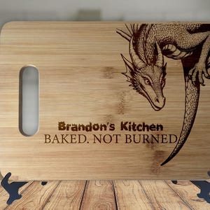 Op de afbeelding: Een rechthoekige bamboe snijplank met een handgreepuitsparing. Een gedetailleerde drakenillustratie is aan de rechterkant geëtst. De tekst "Brandon's Kitchen BAKED. NOT BURNED" staat eronder.