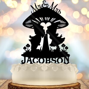May include: A black silhouette cake topper with a mushroom design, featuring the words "Mr & Mrs" at the top. The design includes two figures kissing under the mushroom, with the surname "Jacobson" below. The cake is white with a decorative edge.