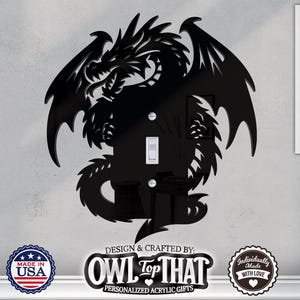 Può includere: Copri interruttore a forma di drago nero. Il drago ha ali dettagliate e una lunga coda. La copertura è progettata per adattarsi a un interruttore della luce standard. Il testo "OWL Top That" e "Made in USA" è visibile.