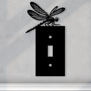 Puede incluir: Tapa de interruptor de luz negra con un diseño de libélula. La libélula está posada en la parte superior de la tapa del interruptor y tiene detalles intrincados.