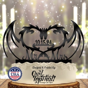 Puede incluir: Topper de pastel de boda negro con dos alas de dragón que forman un corazón, con las palabras "Mr. & Mrs." y la fecha "10.15.24" debajo. El topper está sobre un pastel blanco con una pegatina "Made in USA" roja, blanca y azul.