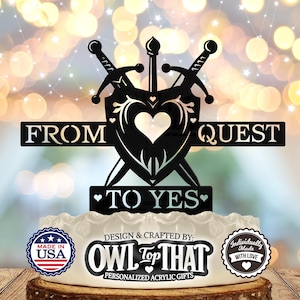 以下が含まれることがあります： 黒い金属製のウェディングケーキトッパー。2本の剣がハート型の盾の後ろで交差している。トッパーには「From Quest to Yes」の文字が書かれている。トッパーは白いケーキの上に置かれており、「Made in USA」のステッカーと「Owl Top That」のロゴが貼られている。