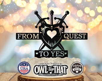 May include: Black metal wedding cake topper with two swords crossed behind a shield with a heart. The text "From Quest to Yes" is on the topper. The topper is on a white cake with a "Made in USA" sticker and a "Owl Top That" logo.